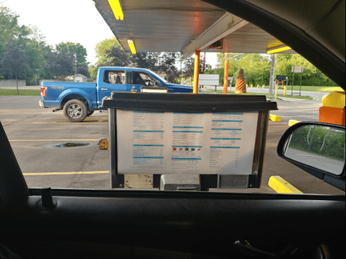A drive-thru menu board is visible, with a blue truck parked nearby in a sunny parking lot.