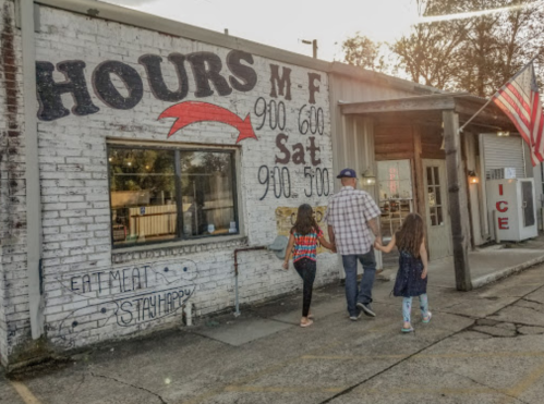 A family walks hand-in-hand past a building with business hours and an American flag. The scene is warm and inviting.