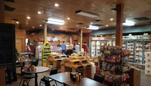Interior of a rustic store with shelves of snacks, a deli counter, and tables, bustling with customers and staff.