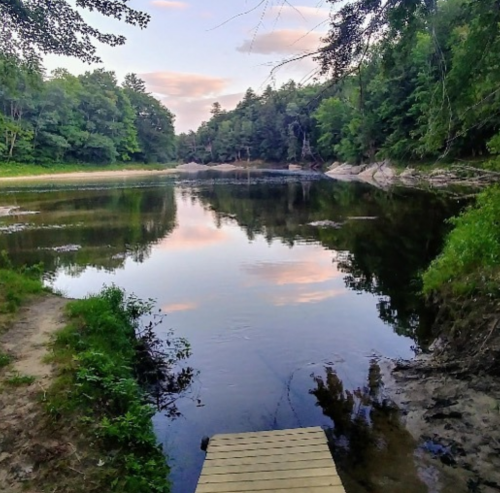 A serene river scene with trees lining the banks and a wooden platform extending over calm water reflecting the sky.