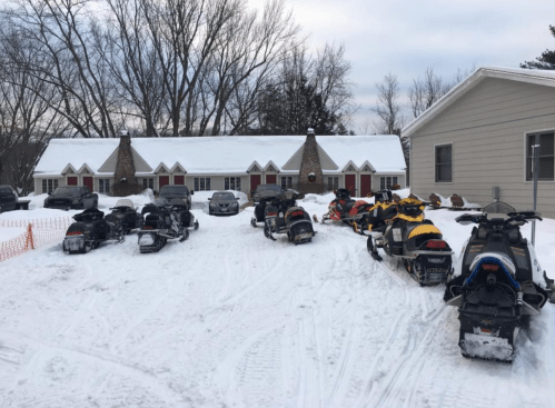 A snowy scene featuring several parked snowmobiles in front of a lodge with stone accents and red doors.