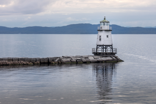A white lighthouse stands on a rocky pier, reflecting in calm waters with mountains in the background under a cloudy sky.