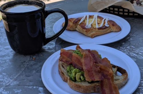 A plate with avocado toast topped with bacon, alongside a pastry drizzled with icing, and a cup of coffee.