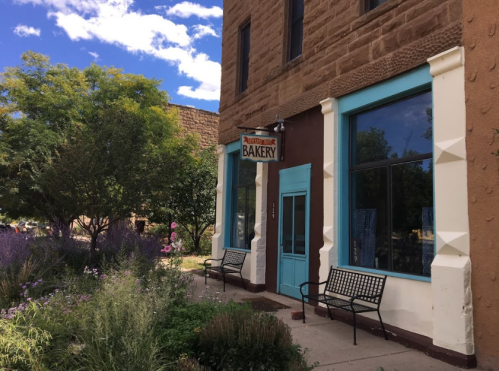 A charming bakery with a turquoise door, surrounded by greenery and flowers, under a blue sky with fluffy clouds.