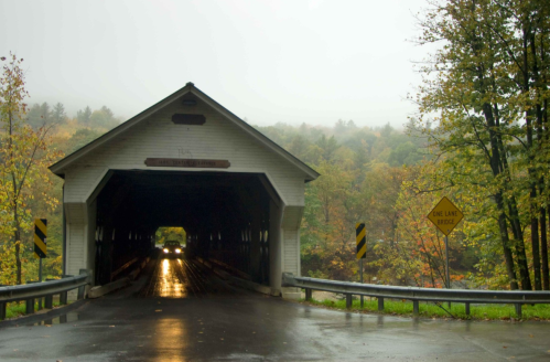 A covered bridge in a misty landscape, with autumn foliage and a sign indicating a one-lane bridge ahead.