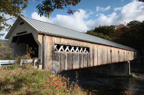 A rustic covered bridge with a wooden exterior spans a river, surrounded by autumn foliage and a cloudy sky.