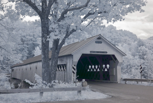 A historic covered bridge surrounded by trees, with a gravel road leading to its entrance.