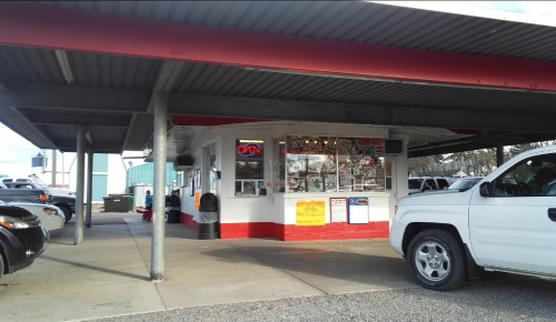 A small food stand with an "Open" sign, surrounded by parked cars under a metal roof.
