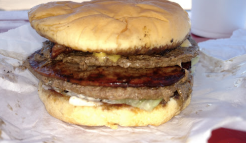 A stacked burger with two beef patties, bacon, pickles, and mayonnaise on a soft bun, resting on a piece of paper.