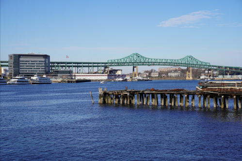 A view of a green bridge over a blue river, with boats and a wooden pier in the foreground under a clear sky.