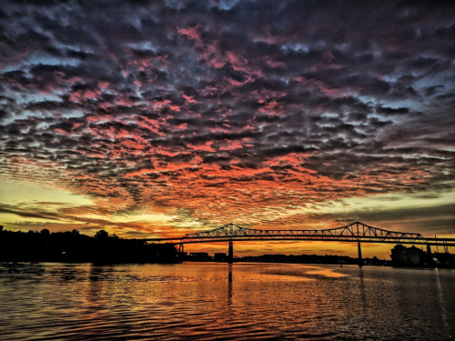 A vibrant sunset over a river, with dramatic clouds and a bridge silhouetted against the colorful sky.