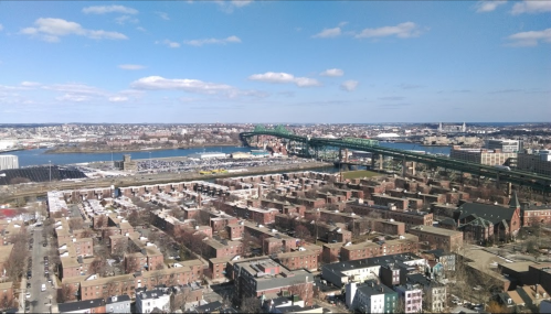 Aerial view of a cityscape featuring buildings, a bridge, and a river under a clear blue sky with scattered clouds.