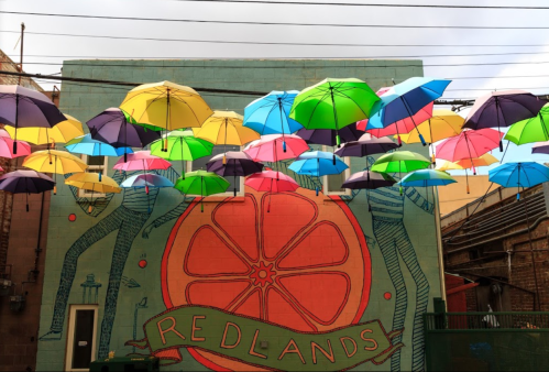 Colorful umbrellas suspended above a mural featuring a large orange and the word "REDLANDS" on a building wall.