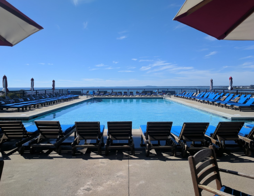 A sunny pool area with blue lounge chairs and umbrellas, overlooking a calm body of water and clear blue sky.