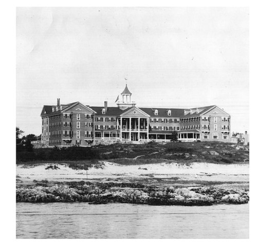 Historic black-and-white photo of a large, multi-story building by the shore, surrounded by rocky terrain.