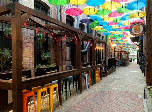 Colorful umbrellas hang overhead in a vibrant alley with wooden seating and plants, creating a lively outdoor dining space.
