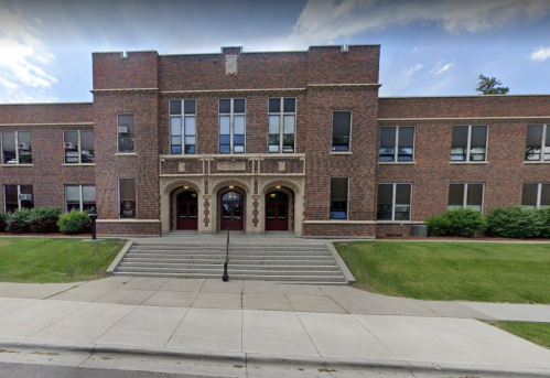 Brick school building with a central entrance, steps, and large windows, surrounded by green grass and trees.