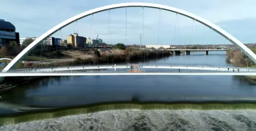 A modern arch bridge spans a river, with a city skyline and a waterfall visible in the background.