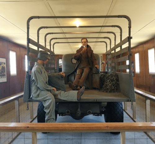 Two life-sized figures in a vintage truck, one in military attire and the other in civilian clothes, inside a museum setting.