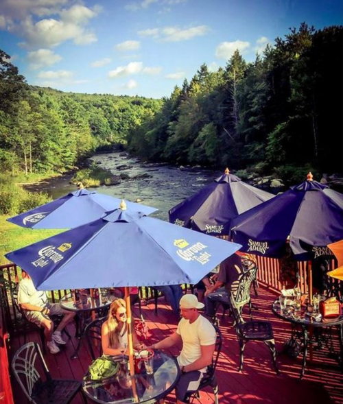 A scenic outdoor dining area by a river, featuring umbrellas and people enjoying drinks in a lush, green setting.