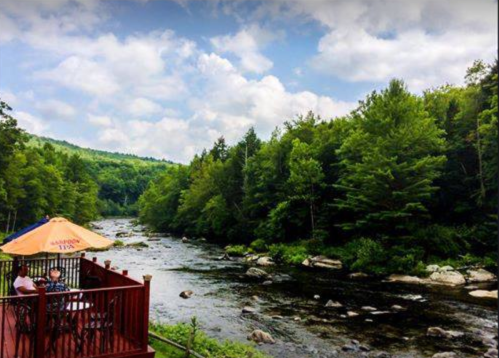 A scenic view of a river surrounded by lush green trees, with a deck and umbrella in the foreground.