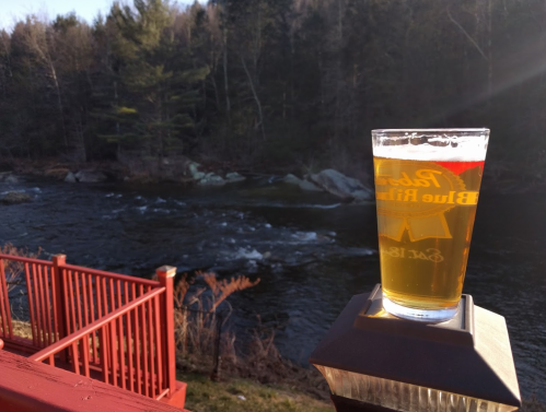 A glass of beer sits on a railing, overlooking a flowing river and wooded landscape in the background.