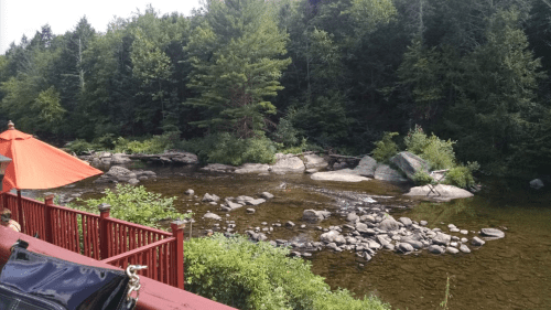 A serene river scene surrounded by lush greenery and rocks, with a glimpse of a red railing and an orange umbrella.
