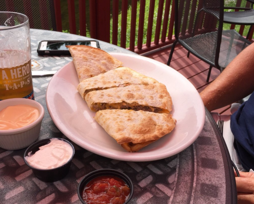 A plate with a large quesadilla cut into slices, accompanied by dipping sauces and a drink in the background.