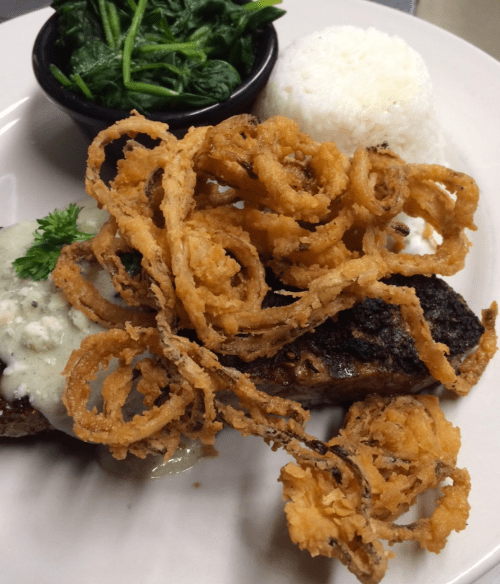 A plate featuring grilled meat topped with creamy sauce, crispy onion rings, steamed spinach, and a serving of rice.