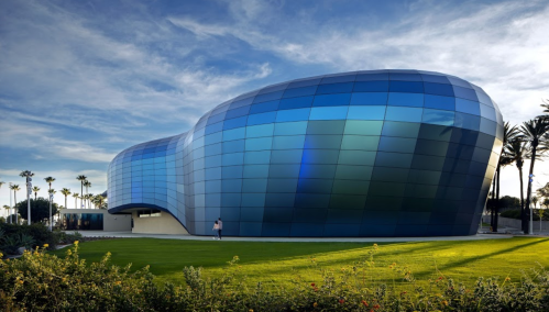 A modern, curvy building with blue glass panels, surrounded by green grass and palm trees under a clear sky.