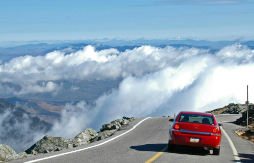 A red car drives along a winding road above the clouds, with mountains visible in the distance under a clear blue sky.