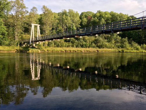 A wooden bridge spans a calm river, reflecting trees and the sky in the water below.