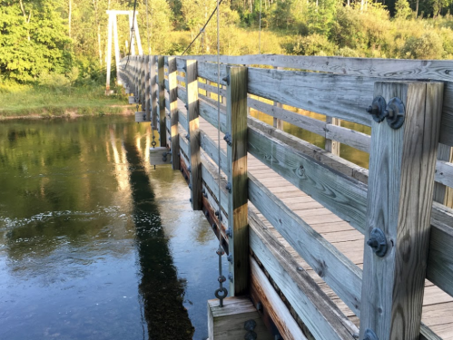 A wooden suspension bridge spans a calm river, surrounded by lush greenery and trees.