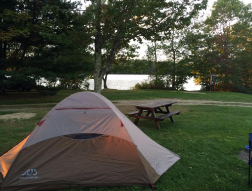 A camping scene featuring a tent by a lake, surrounded by trees and a picnic table in the foreground.