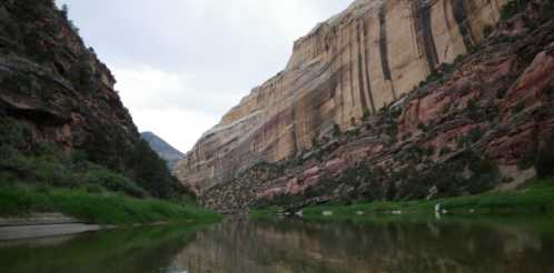 A serene river flows between towering cliffs, surrounded by lush greenery and a cloudy sky.