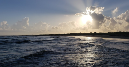 Sunset over a calm ocean, with waves gently lapping at the shore and clouds illuminated by the sun's rays.