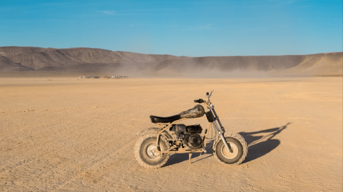A small dirt bike stands alone on a vast, sandy landscape with distant mountains and a clear blue sky.