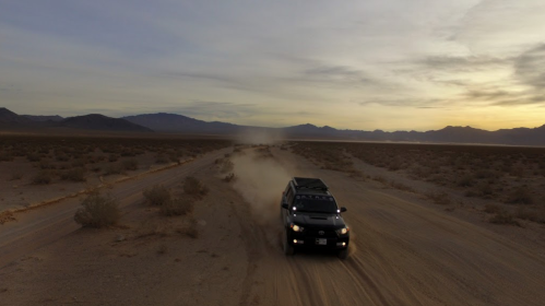 A vehicle drives on a dusty desert road at sunset, with mountains in the background and a clear sky.
