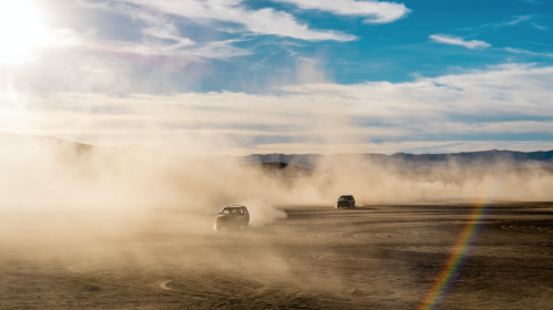 Two cars kick up dust while driving on a barren landscape under a bright sky with wispy clouds and a faint rainbow.