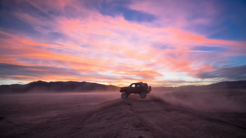 A silhouette of a vehicle jumping over a dusty terrain against a vibrant sunset sky with colorful clouds.