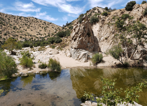 A serene landscape featuring a calm pond surrounded by rocky hills and greenery under a blue sky with scattered clouds.