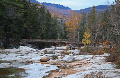 A serene landscape featuring a wooden bridge over a rocky river, surrounded by autumn foliage and mountains.