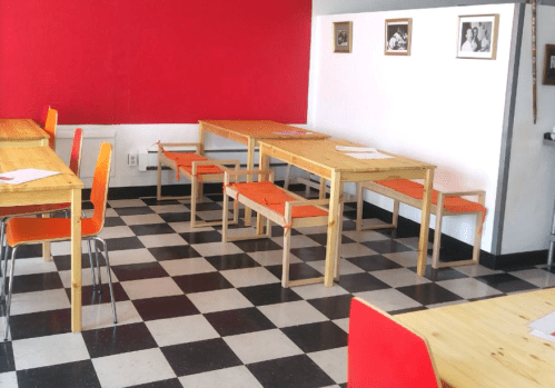 A bright, modern dining area with wooden tables, orange chairs, and a red accent wall. Black and white checkered floor.