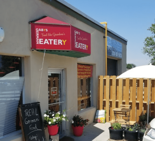 Exterior of Gaby's German Eatery with a red sign, potted plants, and a wooden fence. A chalkboard advertises "Real German Food."