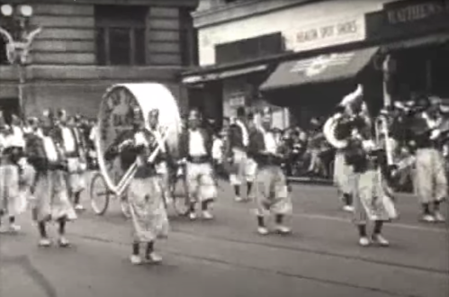 A black and white image of a parade with musicians in traditional attire, featuring a large drum and brass instruments.