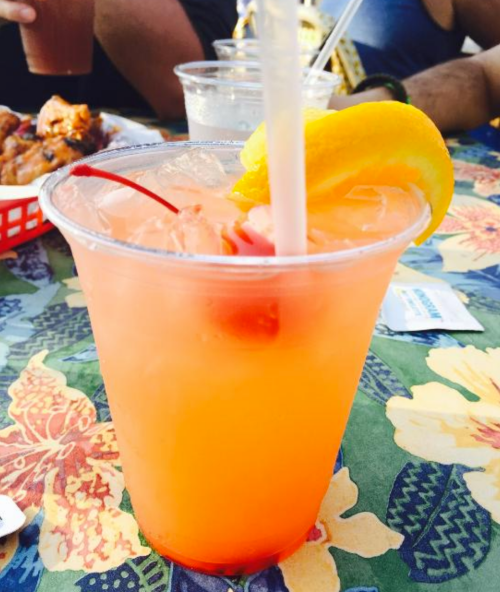 A colorful cocktail with a cherry and lemon slice, served in a clear cup on a floral tablecloth.