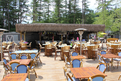 A tropical-themed outdoor dining area with wooden tables and chairs, surrounded by trees and a thatched-roof bar.