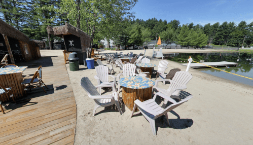 A sandy area with colorful tables and chairs by a calm lake, surrounded by trees and a small building in the background.