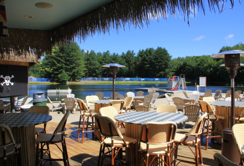 A lakeside patio with tables and chairs, surrounded by trees and a clear blue sky. A dock is visible in the background.