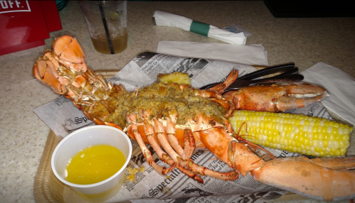 A cooked lobster with stuffing, served with corn on the cob and a cup of melted butter on a tray.